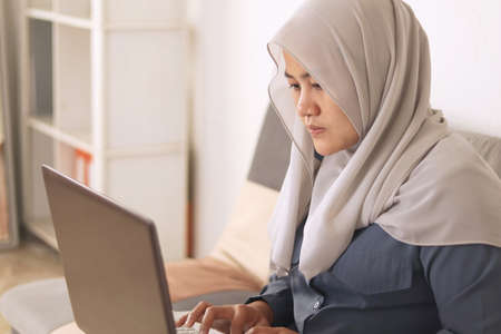Asian muslim woman wearing hijab smiling while typing on her laptop ...