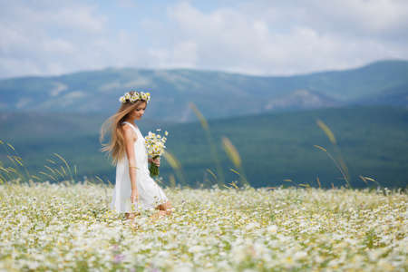 Young beautiful woman brunette with long straight hair, holding a bouquet of beautiful flowers field daisies, dressed in a white sleeveless dress, her head wears a wreath of white flowers field of daisies, walking alone in a flowery field in summer