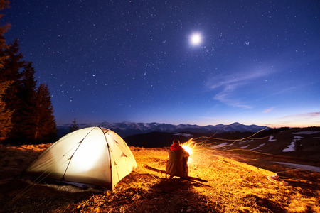 Male tourist have a rest in his camp at night near campfire and tent ...