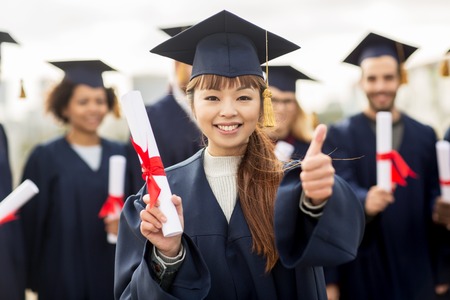 education, graduation, gesture and people concept - group of happy international students in mortar boards and bachelor gowns with diplomas showing thumbs up