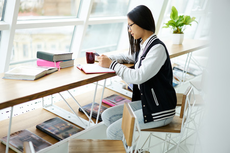 Young chinese international students doing homework tasks in modern university library sitting near window with textbooks and stuff