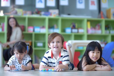 group of international kids in preschool enjoy reading books with teacher watching in back ground