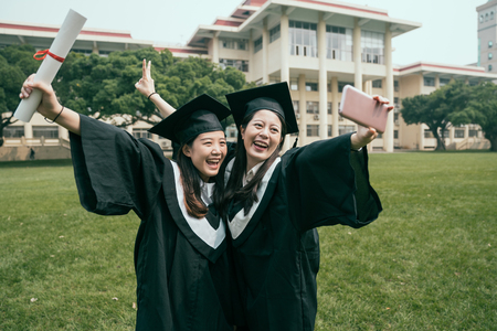 education graduation technology and people concept. group of happy international students in mortar boards and bachelor gowns with diplomas taking selfie by smartphone outdoors on summer sunny day.