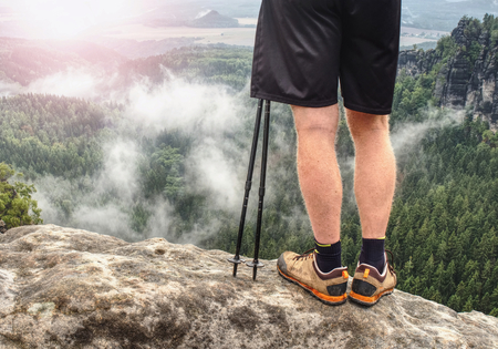 Nordic walking sticks and naked legs in sport shorts. Body of a mountain hiker with hiking boots on a rock.