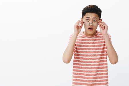 Portrait Of Surprised And Amazed Young Cute Asian Male Student Taking Off Glasses As Hearing Impressive Unexpected News Open Mouth From Surprise Listening To Last Rumors Excited Over White Background Stock