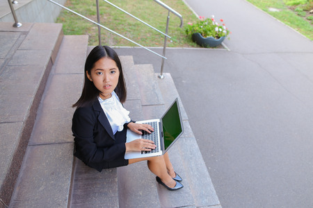 Modern young women, business people, international students holds laptop and study work plan, solve important problems and smiling sits on stairs on background of gray wall of business center outdoors. Woman with black hair, dressed in white blouse and cl