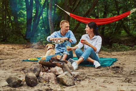 Cheerful couple with dog relaxing near fire in nature - Stock Image ...
