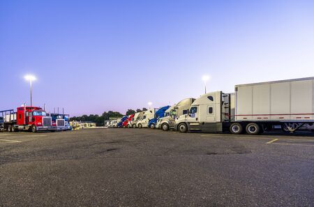 Different make and models big rigs semi trucks with semi trailers standing in row on truck stop parking lot for rest and comply with the movement according to the schedule for successful delivery