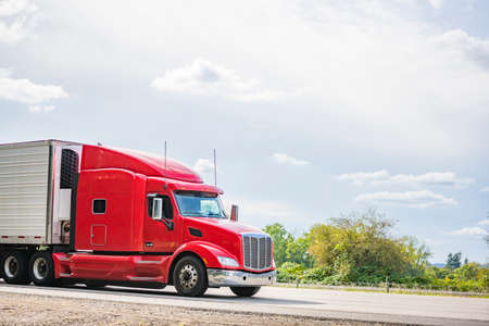 Bright red long hauler big rig red industrial semi truck transporting frozen and chilled foods in refrigerator semi trailer running on the straight highway road with green trees on the side