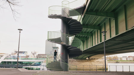 A man stands on a spiral staircase below a bridge, overlooking the smooth water and city buildings.の写真素材