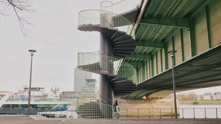 A modern spiral staircase is located under a bridge by the river. Boats are parked nearby, creating a scenic view.の写真素材