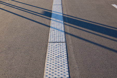 A metal tactile paving strip runs along the road, providing guidance for visually impaired individuals. The sun casts shadows on the textured surface in Neuss, Germany.の写真素材