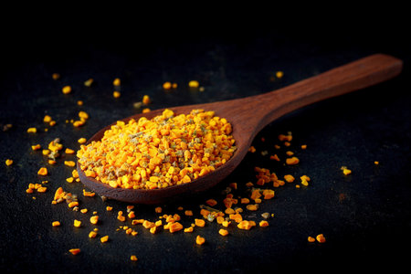 Golden pollen granules rest on a wooden spoon, illuminated by soft studio lighting against a dark background.の素材