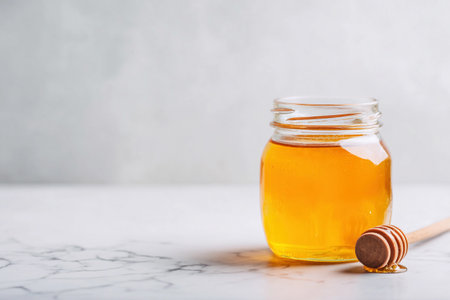 A jar of golden raw honey sits on a marble surface with a dipper placed beside it.の素材
