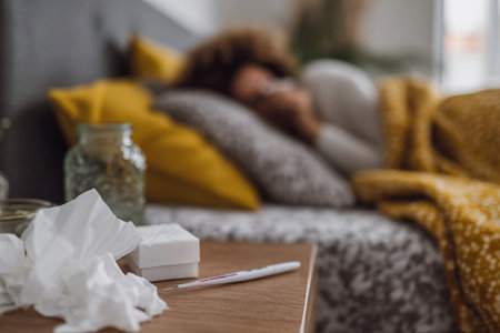 A young woman lies in bed, appearing sick and tired, with tissues and medication nearby.の素材