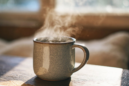 A warm drink sits steaming in a ceramic mug placed on a wooden table near a sunlit window.の素材