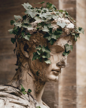 Statue of Greek design shows ivy and roses growing on it in a garden with ruins nearby.の素材