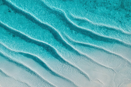 Shallow turquoise water shows clear lines formed by waves as the sand meets the water at noon.の素材