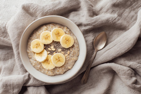Bowl of oatmeal features banana slices and seeds, placed on a cloth background for breakfast.の素材