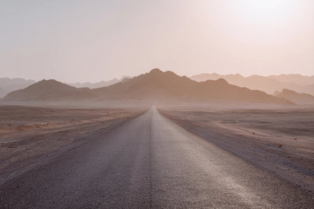 A long road leads through a desert landscape as the sun rises, casting soft light on the scenery.の素材