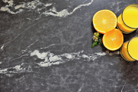 Two glasses of fresh orange juice sit on a dark marble countertop with orange slices nearby.の素材