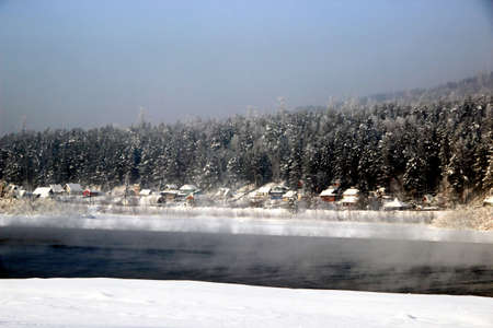 In the background, on the other bank of the river, conifer winter forest begins. Several huts with snow covered roofs near the forest. In the foreground - mist rising from the water and fluffy snow.の写真素材