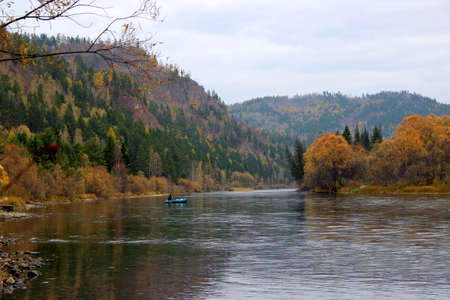 A fast mountain river flows between wooded hills and a lone fisherman on a blue boat. Autumn landscape.の写真素材