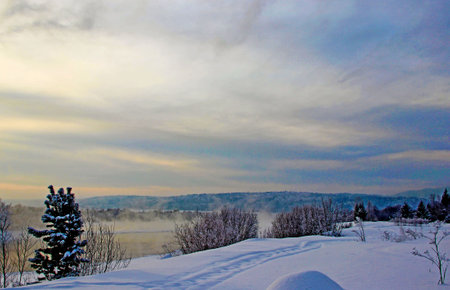 View on a snow covered river bank and wooded mountains in the twilight.の写真素材