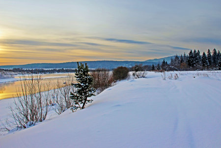 View on a snow covered river bank and wooded mountains in the twilight.の写真素材