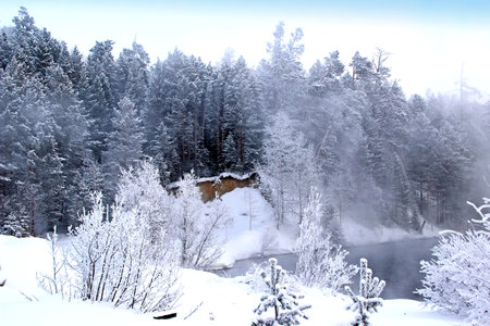 Steep snow-covered river banks are covered with coniferous forest. Clear frosty sky. Fog rises from the water.の写真素材