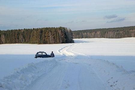 The snow-covered winding road disappears into the forest. The man near the car looks into the distance.の写真素材