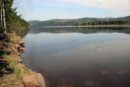 View on a calm water surface of the wild river runs between wooded mountainsの写真素材