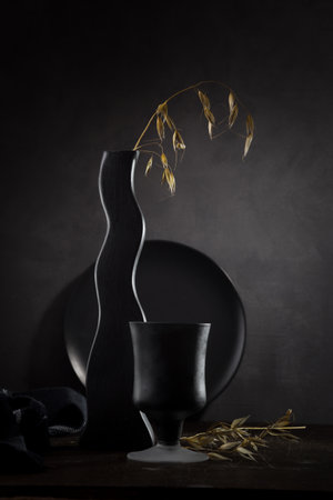 Contemporary still life with dry oat branch in black vase and black dishes on dark background. Black on black.の写真素材
