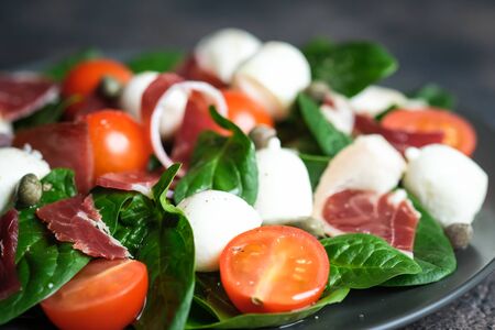 Salad with prosciutto, mozzarella, cherry tomatoes, capers and spinach on a dark plate on a dark background top view copy space.の写真素材