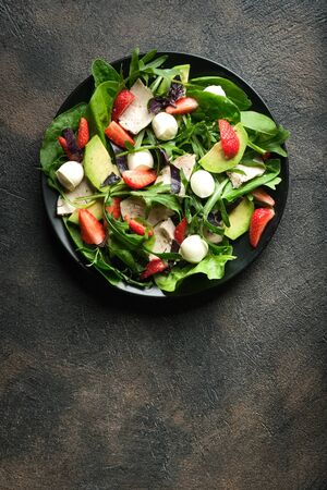 Salad with fresh strawberries, arugula, basil, avocado and mozzarella on a dark background. Healthy vegan breakfast. Top view. Copy space.の写真素材
