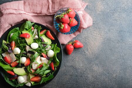 Salad with fresh strawberries, arugula, basil, avocado and mozzarella on a dark background. Healthy vegan breakfast. Top view. Copy space.の写真素材