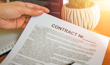 Close-up of a womans hand holding a paper business contract, office background, glow on the background of a laptop, flower, tableの写真素材