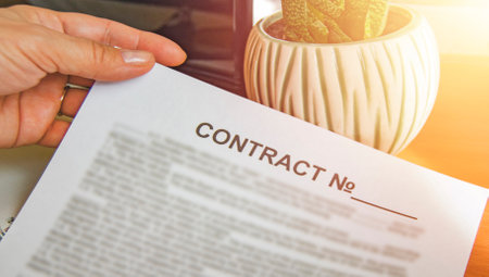 Close-up of a womans hand holding a paper business contract, office background, glow on the background of a laptop, flower, tableの写真素材