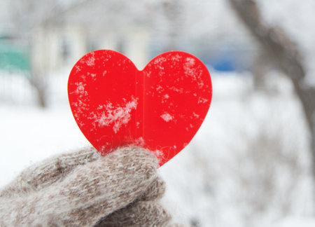 A womans hand in a knitted warm glove holds a red heart, on a blurred background of a city Park, snow, winter, Valentines day concept, close-upの写真素材