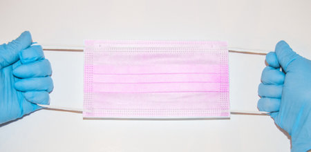 The hands of a medic in blue latex gloves hold a surgical mask with an ear loop on a white...