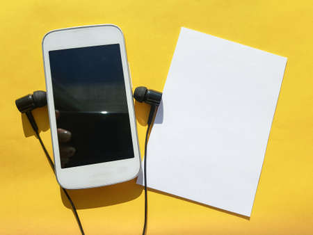 A new concept of using the modern social app Clubhouse on a smartphone, headphones and a blank sheet with a copy of the space on a yellow backgroundの写真素材