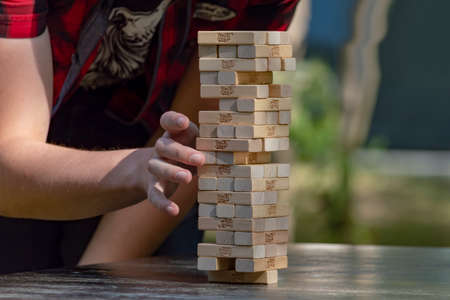 Guy sticks out wooden block from tower of game jenga. Side front view. Concept. Surgut, Russia - September 25, 2019.のeditorial素材