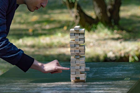 Guy sticks out wooden block from tower of game jenga. Side view. Concept. Surgut, Russia - September 25, 2019.のeditorial素材