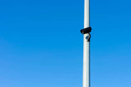 Black CCTV camera on gray pole against blue sky. Sunny winter day. Side view.
