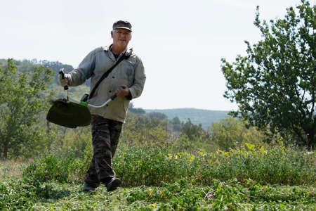 An old man mows grass with gasoline trimmer. Sunny autumn day. Front view.