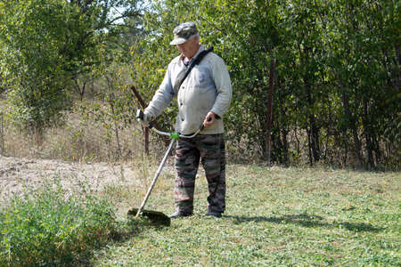 An old man mows grass with gasoline trimmer. Sunny autumn day. Front view.