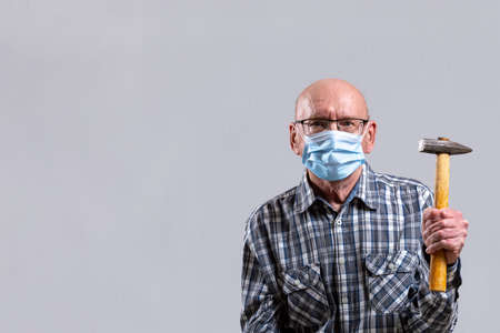 Old bald man with glasses and medical mask with hammer in his hand. Copy space. Front view, indoors vertical shot.の写真素材