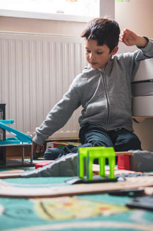 Poznan, Poland - May 16, 2020: Six years old Polish Caucasian boy sitting by a bed and playing with wooden toy train tracks on the floor.のeditorial素材