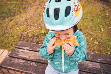 Poznan, Poland - May 17, 2020: Young small boy wearing a safety helmet eating a fresh waffle while sitting on a wooden bench.Â のeditorial素材