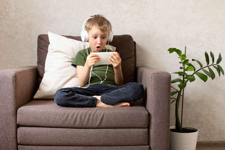 A child boy with a phone sits on a chair with headphones watching cartoons, listening to music, playing games, talking on the phone.の写真素材
